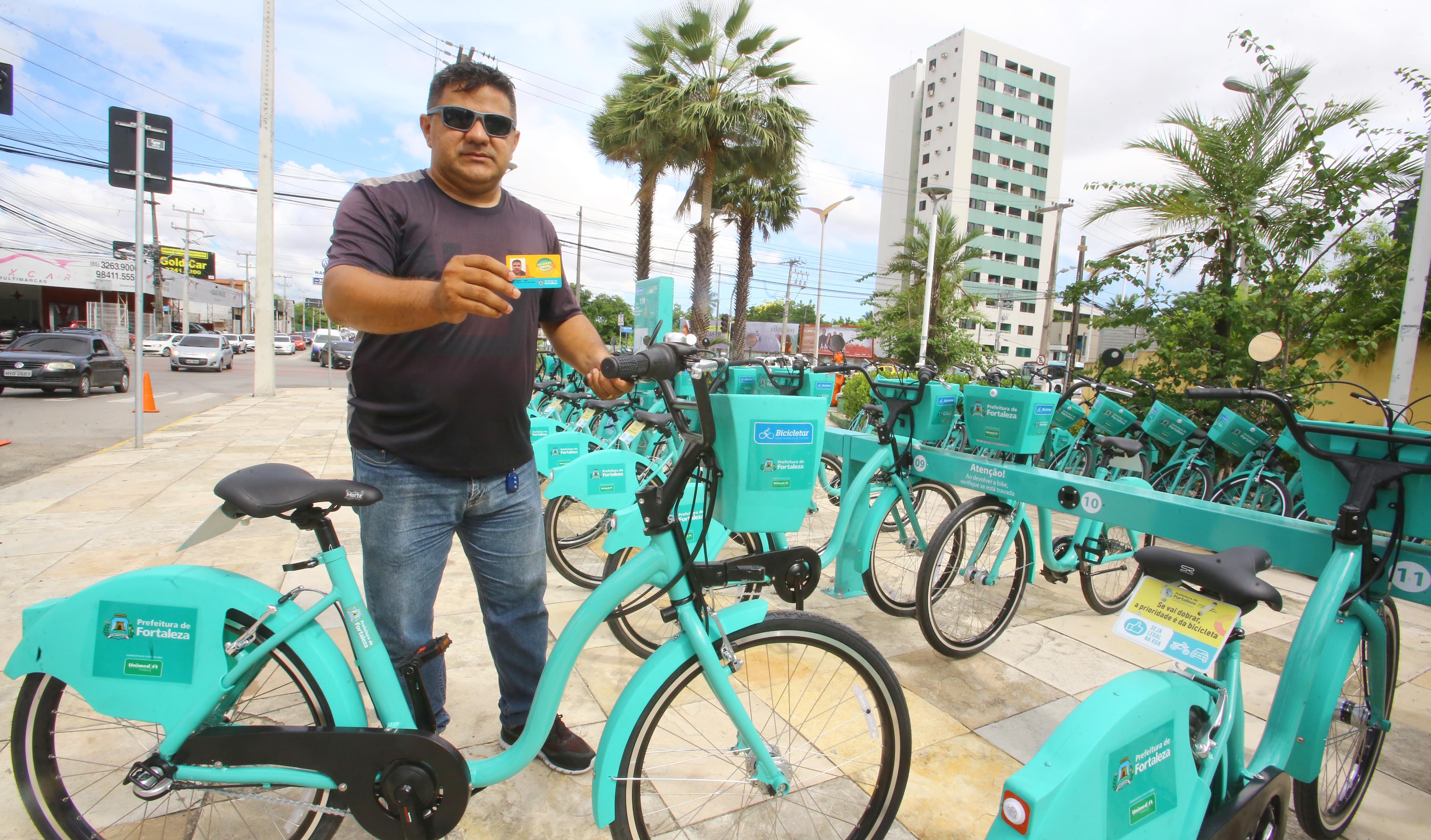 homem de óculos escuro posando para a foto ao lado de estação de bicicletas e segurando bilhete único